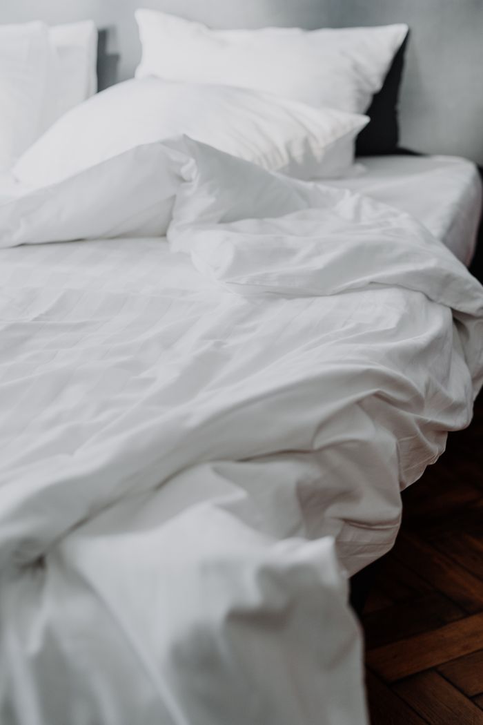 services-01 A close-up of a cozy white linen bed with rumpled bedding in a modern bedroom setting.